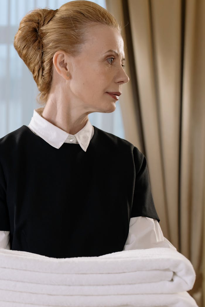 Elegant housekeeper in uniform holding fresh towels in a well-lit hotel room.